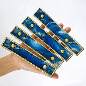 Puede incluir: Cinco piezas decorativas rectangulares con un diseño veteado azul y blanco, enmarcadas en dorado y acentuadas con tachuelas doradas. Las piezas se sostienen en una mano sobre un fondo blanco.