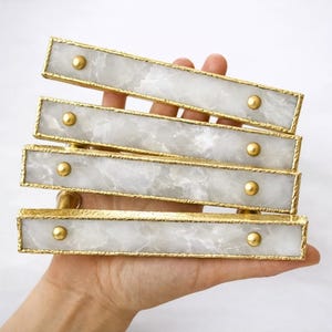 May include: A set of four rectangular cabinet pulls. Each pull features a white marble-like center, framed by gold-colored metal with a textured edge. Gold-colored round studs secure the marble to the frame. The pulls are held in a hand.