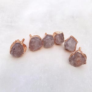 Puede incluir: Seis tiradores de armario de cristal de amatista en bruto con bordes y tallos dorados. Los tiradores son de color púrpura claro y tienen una textura rugosa y natural. Están dispuestos en semicírculo sobre una superficie blanca.