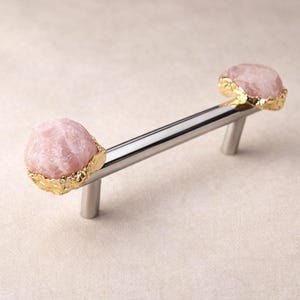 Peut inclure: Une poignée de meuble avec une barre argentée polie et deux extrémités en pierre de quartz rose, chacune bordée d'or. Ce matériel de mobilier allie élégance naturelle et design contemporain.