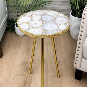 May include: A circular side table featuring a white and grey agate stone top with gold edging. The table is supported by three gold-coloured legs. A decorative accent piece for a living room or bedroom.