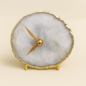 May include: A round, white and gray agate clock with gold-colored hands and a gold-plated edge. The clock has a gold-colored base with three small legs. The clock face has a natural, marbled appearance.