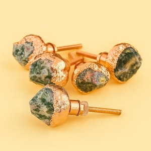 Puede incluir: Seis tiradores decorativos para armarios con aspecto de piedra natural. Los tiradores tienen una base de metal dorado y están parcialmente recubiertos con pan de oro. La piedra tiene tonos verdes y marrones, sobre fondo amarillo.