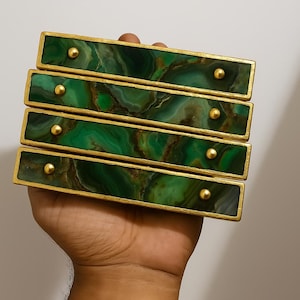May include: Four rectangular drawer pulls with a green and brown marbled pattern, framed in gold, and adorned with gold-colored knobs. The pulls are held in a hand against a neutral background.