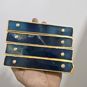 Könnte beinhalten: Vier rechteckige Schubladengriffe mit goldfarbenem Rand und Messingknöpfen. Die Griffe haben ein dunkelblaues und weißes Marmor-Aussehen. Die Griffe werden in einer Hand vor einem weißen Hintergrund gehalten.