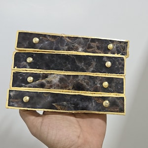 Könnte beinhalten: Vier rechteckige Schubladengriffe mit dunkler, marmorierter Steinoptik, goldfarbenen Zierleisten und kleinen, runden goldenen Knöpfen. Die Griffe sind gestapelt und werden in einer Hand gehalten. Der Hintergrund ist schlicht.