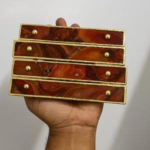 May include: Four rectangular cabinet pulls with a reddish-brown, marbled appearance and gold-colored trim. Each pull has a single, small, gold-colored knob. The pulls are stacked and held in a hand against a white background.