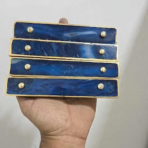Op de afbeelding: Vier rechthoekige decoratieve stukken met een diepblauw, gemarmerd ontwerp en gouden rand. Elk stuk heeft vier kleine, goudkleurige studs. De items zijn gestapeld en worden in een hand gehouden.