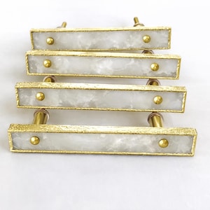 Puede incluir: Cuatro tiradores rectangulares para armarios con un centro blanco similar al mármol y un borde dorado. Cada tirador tiene dos tornillos dorados. Los tiradores están dispuestos en una pila, con el tirador superior ligeramente desplazado.