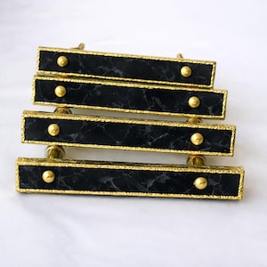 May include: Four rectangular cabinet pulls with black marble-effect centres and gold-coloured trim. Each pull has two gold-coloured, rounded knobs. The pulls are arranged horizontally, suggesting they are for a drawer or cabinet.