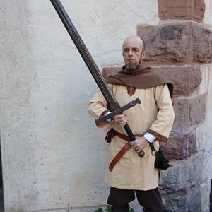 Könnte beinhalten: Ein langes, zweihändiges Schwert mit dunkler, verwitterter Oberfläche. Das Schwert wird von einer Person gehalten, die eine beige Tunika, braune Shorts und schwarze Stiefel trägt. Das Bild enthält das Logo "FENRIS WEAPONRY & PROPS" unten.