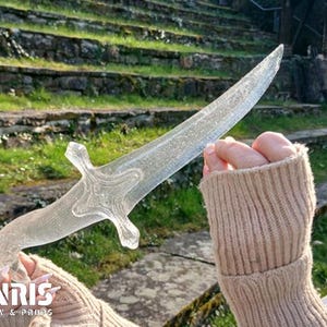 Op de afbeelding: Een heldere, decoratieve dolk met een kruisvormige handgreep wordt omhoog gehouden tegen een achtergrond van stenen trappen en groen. De dolk heeft een getextureerd, kristallijn uiterlijk. Het logo "FENRIS WEAPONRY & PROPS" is zichtbaar.