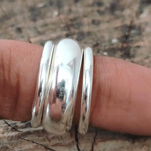 May include: A silver ring with a unique design. The ring features three bands, with the center band being the largest and most prominent. The bands have a smooth, polished finish, reflecting light. The ring is shown on a finger.