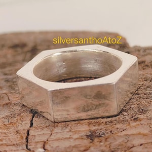 May include: A silver-colored, geometric ring with a hexagonal design. The ring is made of a solid metal and has a smooth, polished surface. The ring is displayed on a wooden surface. The text "silversanthoAtoZ" is in the image.