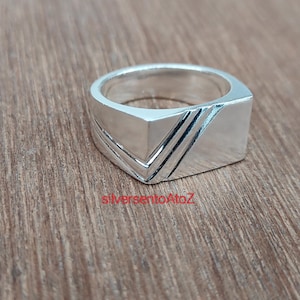 May include: A silver ring with a geometric design. The ring has a rectangular face with a V-shaped design and three parallel lines. The ring is made of a shiny, reflective metal.