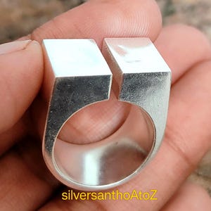 Può includere: Un anello d'argento dal design moderno. L'anello presenta una parte superiore rettangolare con gambo diviso. L'anello è tenuto in una mano. Il testo "silversanthoAtoZ" è visibile sull'anello.