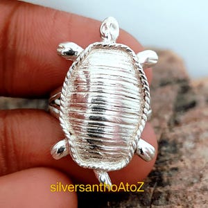 May include: A silver turtle ring with a textured shell design and a twisted rope border. The ring has a turtle head and flipper details. The text "silversanthoAtoZ" is visible in yellow at the bottom of the image.