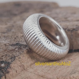 Könnte beinhalten: Ein silberner Ring mit einer strukturierten, geriffelten Oberfläche. Der Ring hat ein dickes Band und ein glattes Inneres. Der Ring wird auf einer Holzoberfläche präsentiert. Der Text "silversanthoAtoZ" ist sichtbar.