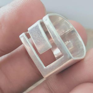 以下が含まれることがあります： 幾何学模様のシルバーカラーのリング。リングは、正方形の開口部と隆起した長方形の要素を備えた長方形のバンドが特徴です。リングは人の指で保持されています。