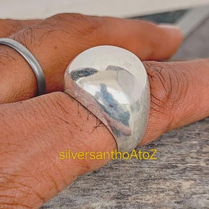 May include: A large, polished silver ring with a rounded, dome-like top. The ring is worn on a finger, and the surface reflects light. The text "silversanthoAtoZ" is visible in the image.