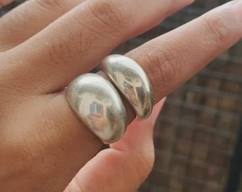 May include: Two silver rings with a smooth, rounded design. The rings are worn on a finger, showcasing their polished, reflective surface. The rings have a modern, minimalist aesthetic.