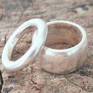 Puede incluir: Dos anillos de plata. Uno es una banda gruesa, el otro es un anillo más pequeño, de forma ovalada. Ambos anillos tienen una superficie pulida y reflectante. Los anillos se muestran sobre una superficie marrón texturizada.