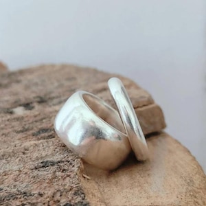 Puede incluir: Dos anillos de plata con un acabado liso y pulido. Un anillo presenta una banda ancha y gruesa, mientras que el otro es una banda fina y delgada. Ambos anillos tienen una superficie metálica reflectante y están colocados juntos.