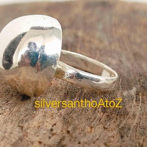 May include: A silver ring with a large, rounded, hammered-texture top. The band is smooth and simple. The ring sits on a rough, brown wooden surface. The text "silversanthoAtoZ" is visible in yellow.