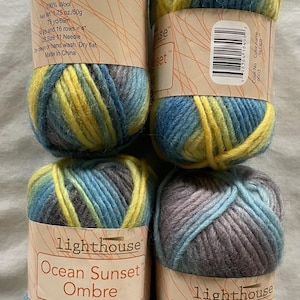 May include: Four skeins of Lighthouse Ocean Sunset Ombre yarn. Each skein is a blend of blue, yellow, and gray wool. The labels indicate the yarn is bulky, 1.75 oz (50g), and 75 yards (69m).