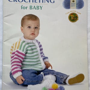 Puede incluir: Un beb&eacute; con un c&aacute;rdigan de ganchillo multicolor y vaqueros est&aacute; sentado frente a un libro titulado "CROCHETING for BABY". La portada del libro presenta el texto "16 ADORABLE BABY ACCESSORIES" y una madeja de lana azul.
