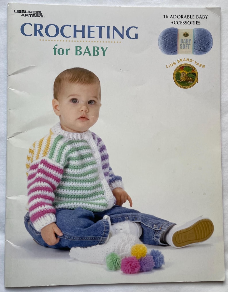 Puede incluir: Un beb&eacute; con un c&aacute;rdigan de ganchillo multicolor y vaqueros est&aacute; sentado frente a un libro titulado "CROCHETING for BABY". La portada del libro presenta el texto "16 ADORABLE BABY ACCESSORIES" y una madeja de lana azul.