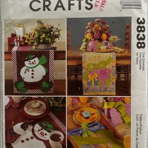 May include: A sewing pattern for a table runner with a snowman design. The pattern includes instructions for making a table runner with a snowman design, a table runner with a gift box design, and a table runner with a striped design. The pattern is from McCall's Crafts and is called "One Envelope - Un Sobre - Pattern/Patron Cont. Un Patrone de Costura".