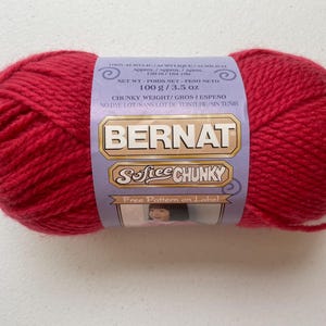 May include: A skein of bright red Bernat Softee Chunky yarn with a light purple paper label. The label displays the text Bernat Softee Chunky, 100 g / 3.5 oz, 150 m / 164 yds, and indicates that it is 100% acrylic with no dye lot.