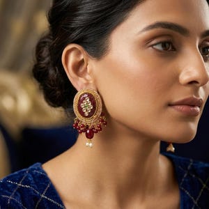 May include: Gold-toned oval earrings with a maroon center and a green and white design. The earrings have dangling red beads and small white pearls. The woman in the image is wearing a blue velvet top.