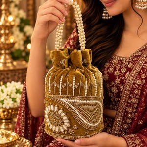 May include: A mustard yellow velvet drawstring handbag featuring intricate white pearl beadwork, gold embroidery, and a paisley motif. The bag has a handle made of multiple strands of white pearls and a gathered top closure with a pearl accent.