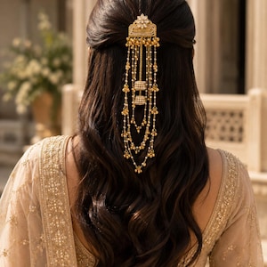 May include: A gold hair accessory with dangling chains and pearl accents is set against dark brown hair. The ornate piece is attached to the hair with a decorative floral design. The woman is wearing a gold-coloured embroidered top.