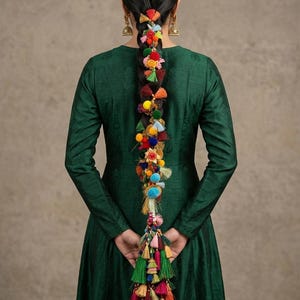 May include: A long, dark braid adorned with colorful pom-poms, tassels, and floral accents. The braid cascades down the back of a person wearing a dark green dress. The decorative elements include red, orange, yellow, blue, and green.