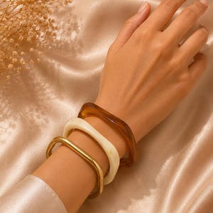 Puede incluir: Un conjunto de tres brazaletes de forma cuadrada que se llevan en la muñeca. La colección incluye un brazalete de metal dorado pulido, un brazalete de resina opaca de color crema liso y un brazalete de resina ámbar translúcido brillante.