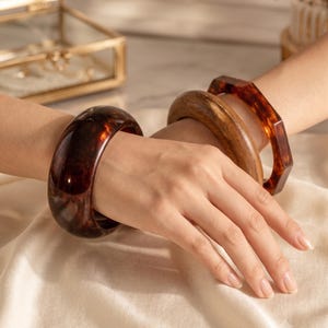 May include: A set of three bangles worn on the wrist. One is a wide, smooth, dark brown tortoiseshell-patterned resin bangle. The second is a smooth, rounded, medium-brown wooden bangle. The third is a thin, dark brown tortoiseshell-patterned resin bangle.