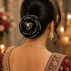 Könnte beinhalten: Ein schwarzes Haarknoten-Accessoire mit einem kreisförmigen Blumenmuster, das mit goldfarbenen Perlen und kleinen klaren Kristallen verziert ist. Das Accessoire sitzt mittig auf einem dunklen Haarknoten, ergänzt durch goldfarbene Ohrhänger und eine feine Kette.