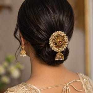 May include: A gold-toned hair accessory featuring a circular filigree design adorned with small white pearls, red and green stones, and a dangling bell-shaped charm. A matching bell-shaped earring with white pearl accents is visible on the ear.