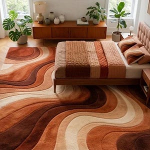 A rectangular rug with wavy stripes in shades of brown, orange, and cream on a light wood floor, extending under a bed with a striped brown and orange comforter.