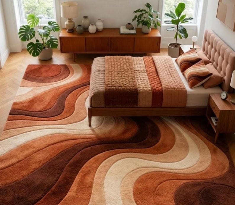 A rectangular rug with wavy stripes in shades of brown, orange, and cream on a light wood floor, extending under a bed with a striped brown and orange comforter.