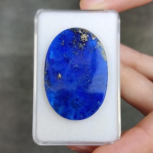 Puede incluir: Gema ovalada de lapislázuli en una funda de plástico transparente. La piedra es de color azul intenso con motas doradas y una superficie pulida. La gema está sobre un fondo blanco.