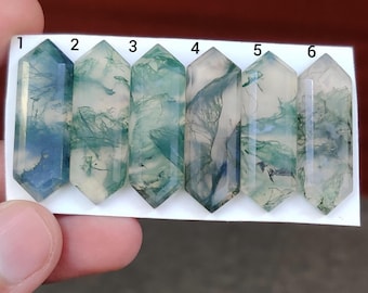 Könnte beinhalten: Sechs klare, sechseckige Moosachat-Kristallstäbe. Die Stäbe zeigen unterschiedliche Muster in Grün, Blau und Weiß, die an Naturlandschaften erinnern. Die Kristalle werden vor einem neutralen Hintergrund gehalten.