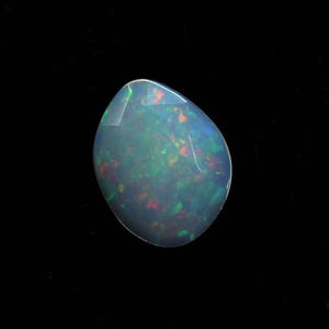 Può includere: Una gemma di opale lucidata a forma di goccia su sfondo nero. L'opale mostra un gioco di colori, con lampi di verde, rosso e blu all'interno del suo corpo traslucido. La superficie della pietra è liscia e riflettente.
