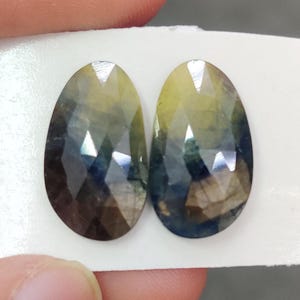 Op de afbeelding: Twee gefacetteerde, ovale edelstenen. De edelstenen hebben een kleurovergang, van geel naar blauw en bruin. De stenen liggen op een witte achtergrond, waardoor hun gepolijste oppervlakken en interne reflecties worden benadrukt. Ideaal voor het maken van sieraden.