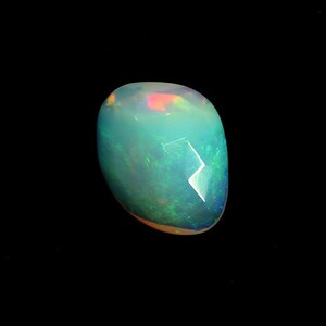 Puede incluir: Una piedra preciosa de ópalo pulida con forma redondeada. El ópalo tiene un aspecto lechoso translúcido con destellos iridiscentes verdes, azules y rojos. La piedra preciosa está sobre un fondo negro.