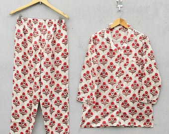 Könnte beinhalten: Ein zweiteiliges Pyjama-Set mit weißem Hintergrund und einem sich wiederholenden roten Blumenmuster. Das Set umfasst ein Hemd mit Knöpfen und eine passende Hose, beide hängen an Holzbügeln.
