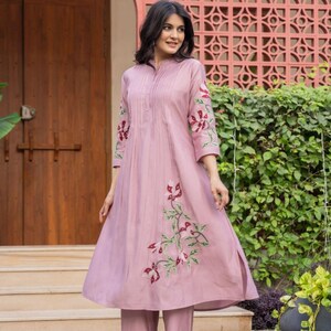 May include: A mauve-colored A-line kurta with three-quarter sleeves and matching straight-leg trousers. The garment features vertical pleats along the chest and embroidered floral motifs in shades of red, white, and green on the sleeves and skirt.